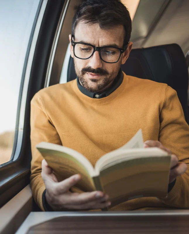 Un uomo è seduto vicino al finestrino di un treno, perso nei suoi pensieri mentre legge un libro
