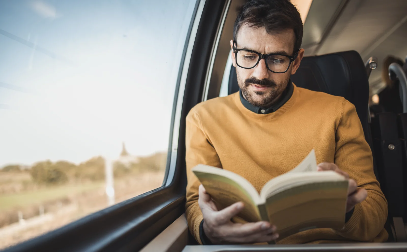 Un uomo è seduto vicino al finestrino di un treno, perso nei suoi pensieri mentre legge un libro